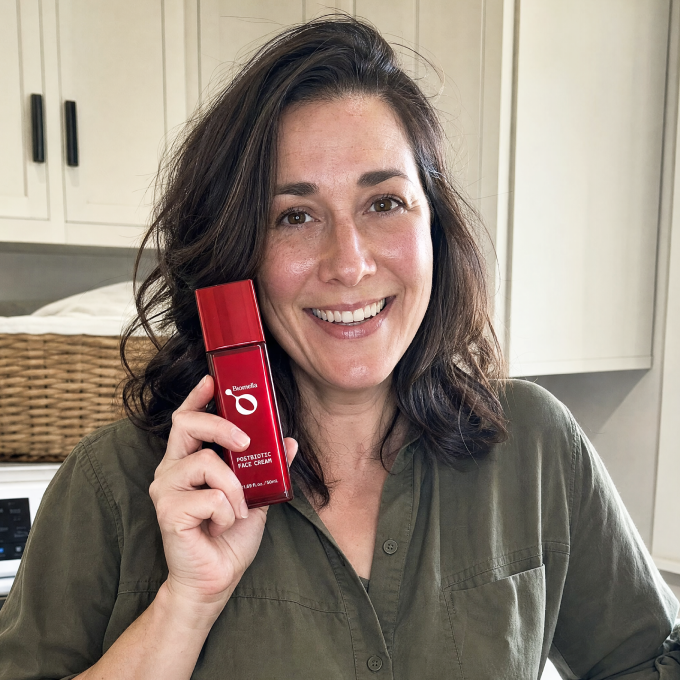 Smiling woman holding Biomella Postbiotic Face Cream red bottle beside her face